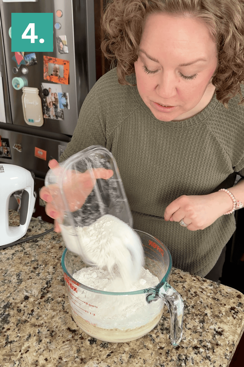 A person pours flour from a container into a large glass measuring cup filled with wet ingredients on a kitchen counter. A white hand mixer is nearby, and a number 4 is in the top left corner.