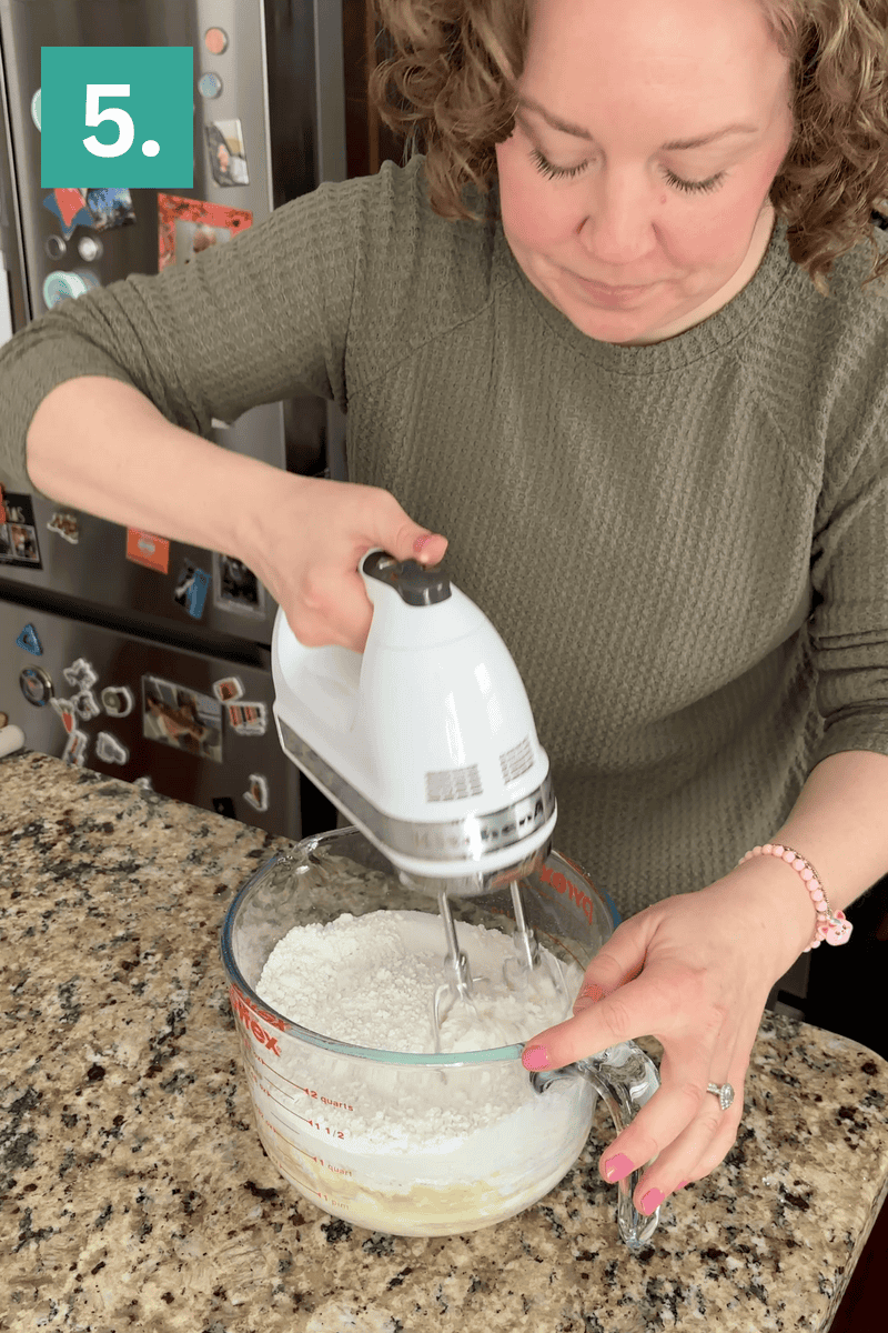 A person in a green sweater uses a hand mixer to blend flour and ingredients in a large glass measuring cup on a granite countertop. A teal box with the number 5 is in the top left corner.