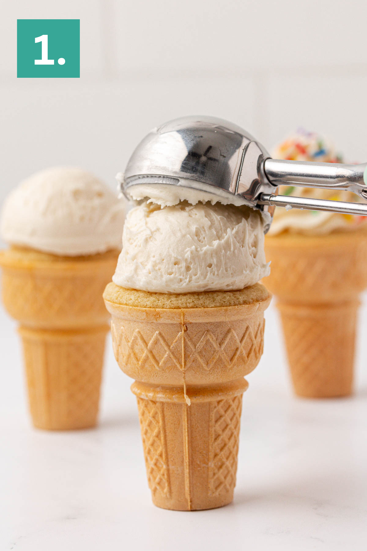 A metal ice cream scoop places a scoop of vanilla ice cream onto an ice cream cone, with two more cones in the background. A green box with the number 1. appears in the top left corner.