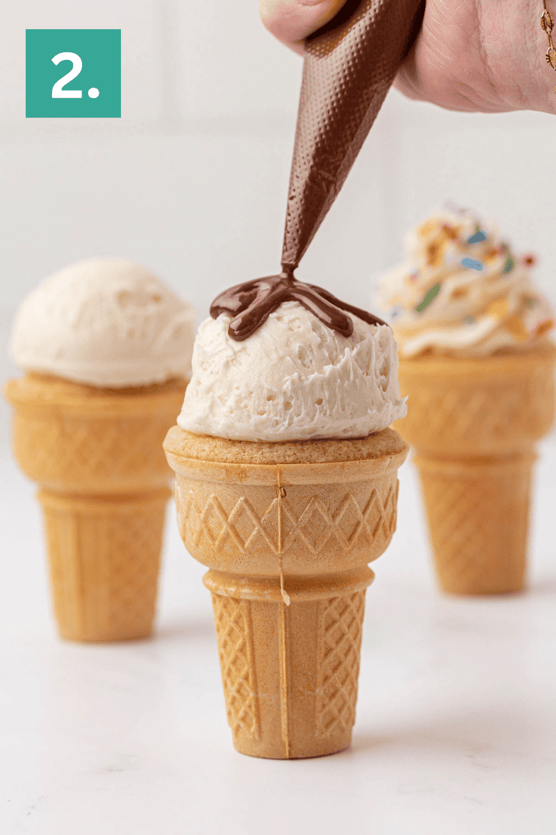 A hand pipes chocolate sauce onto a scoop of vanilla ice cream on a cone-shaped cake, with two more ice cream cone cakes, one plain and one with rainbow sprinkles, in the background. A green box with the number 2 is in the top left corner.