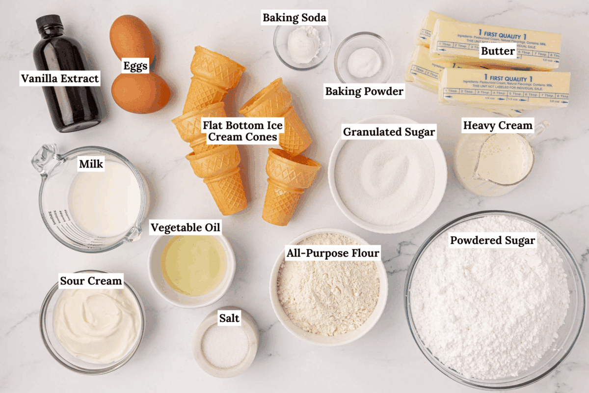 Assorted baking ingredients labeled on a white surface, including eggs, butter, milk, vanilla extract, sour cream, vegetable oil, salt, flour, granulated and powdered sugar, baking soda, baking powder, heavy cream, and ice cream cones.