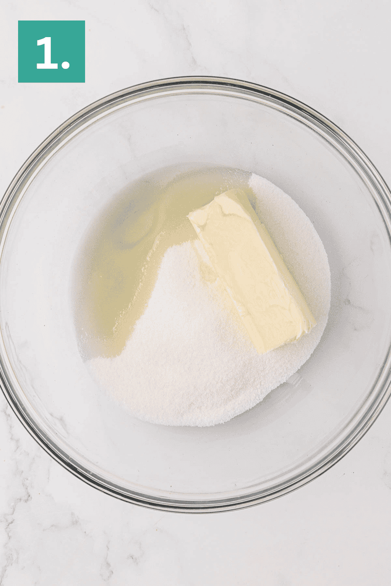 A glass bowl containing a stick of butter, granulated sugar, and egg whites sits on a marble surface. A green square with the number 1. is in the top left corner.
