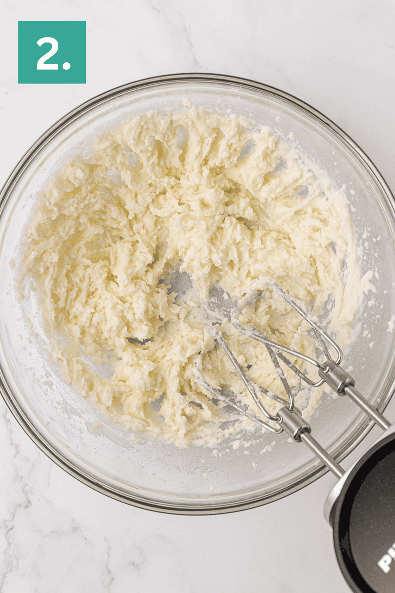 A glass bowl with creamed butter and sugar being mixed by a hand mixer. The number 2. in a green box is in the top left corner, indicating this is the second step of a recipe.