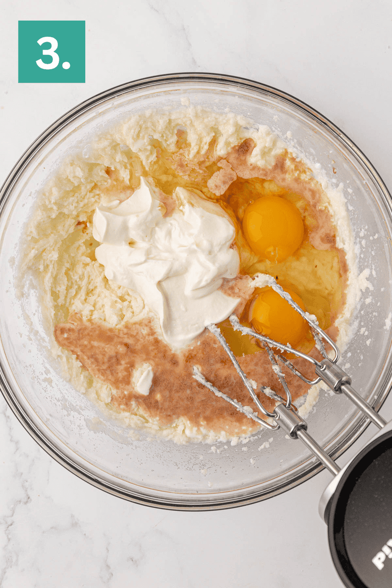 A glass bowl with creamed ingredients, two cracked eggs, sour cream, and vanilla extract, ready to be mixed with electric beaters. A green box labeled 3. is in the top left corner.