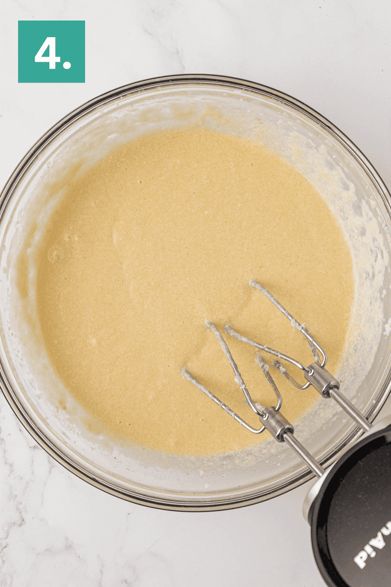 A glass bowl filled with light yellow batter and an electric hand mixer, set on a white marble surface. A green box with the number 4 is in the top left corner.