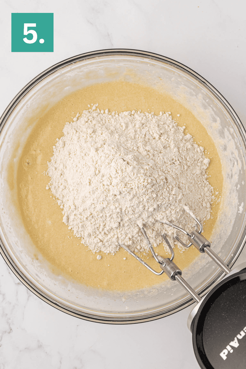 A glass bowl contains a yellow batter with a mound of flour on top. An electric hand mixer with beaters is positioned in the bowl. A green square with the number 5 is in the upper left corner.