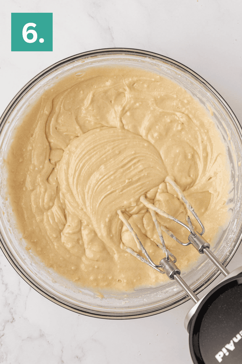 A glass bowl filled with smooth, light yellow cake batter is being mixed with an electric hand mixer. The mixture appears creamy and well-blended. A green box with the number 6 is in the top left corner.