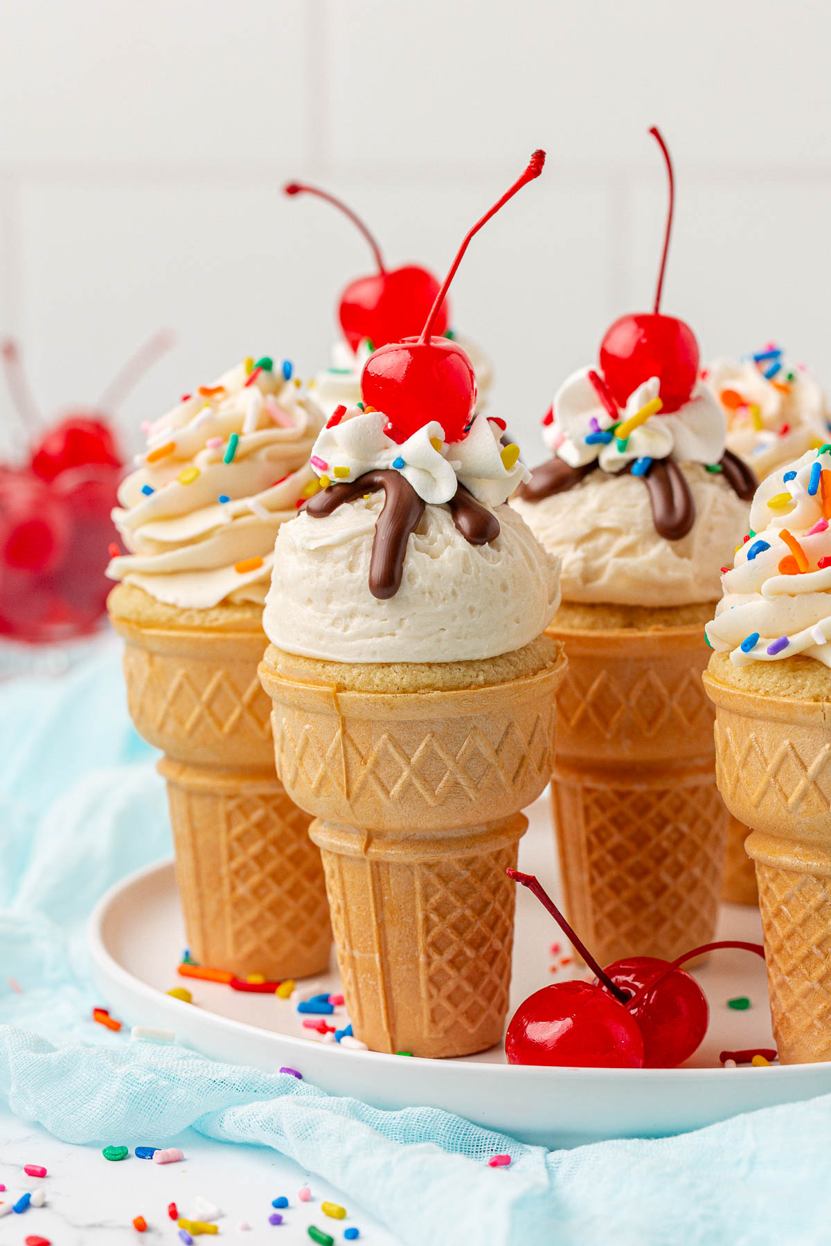 Ice cream cone cupcakes topped with swirls of frosting, colorful sprinkles, drizzled chocolate, and a cherry on top, arranged on a white plate with extra cherries and sprinkles scattered around.