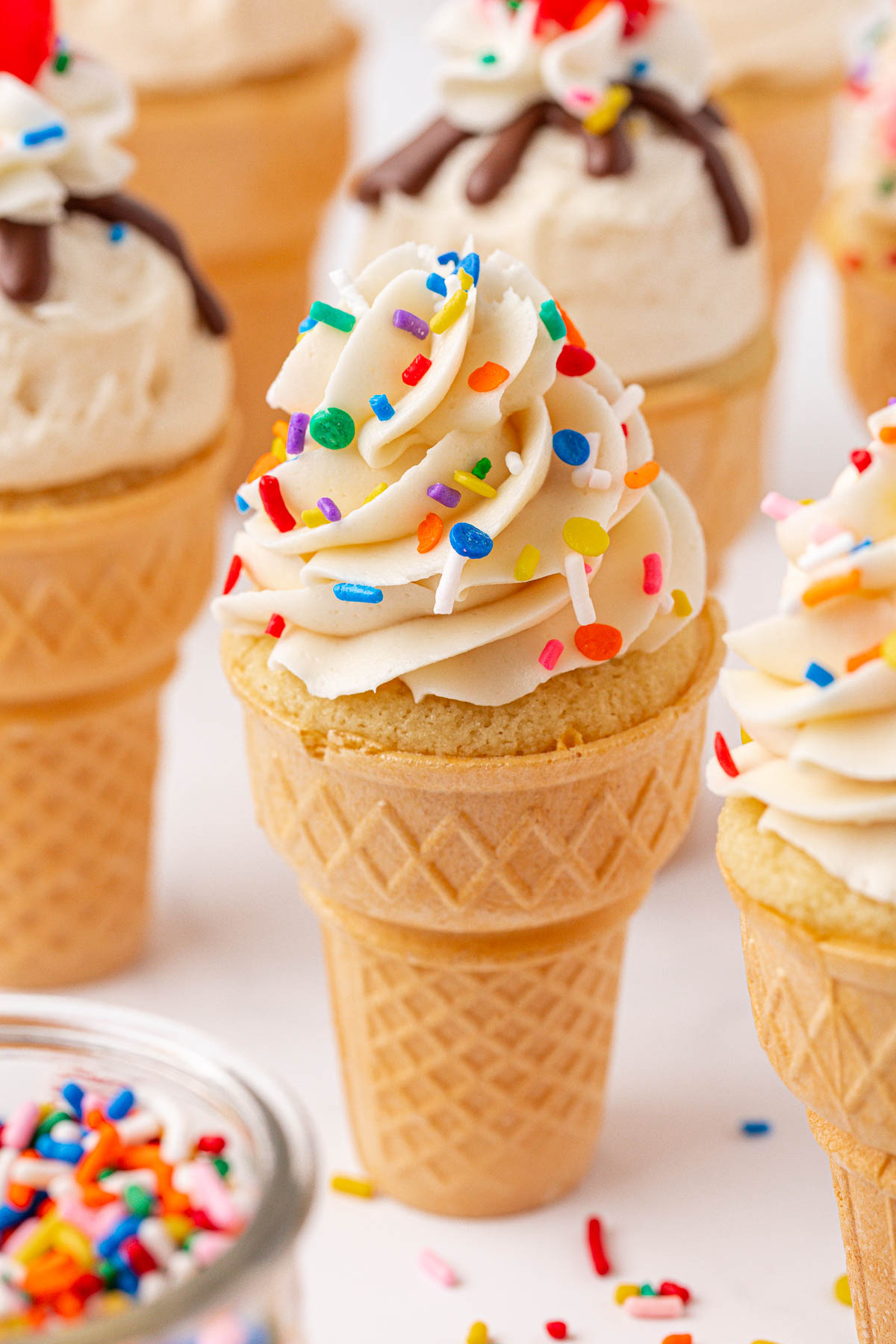 A cupcake baked in an ice cream cone, topped with swirled white frosting and colorful sprinkles, surrounded by similar decorated treats and a jar of sprinkles.