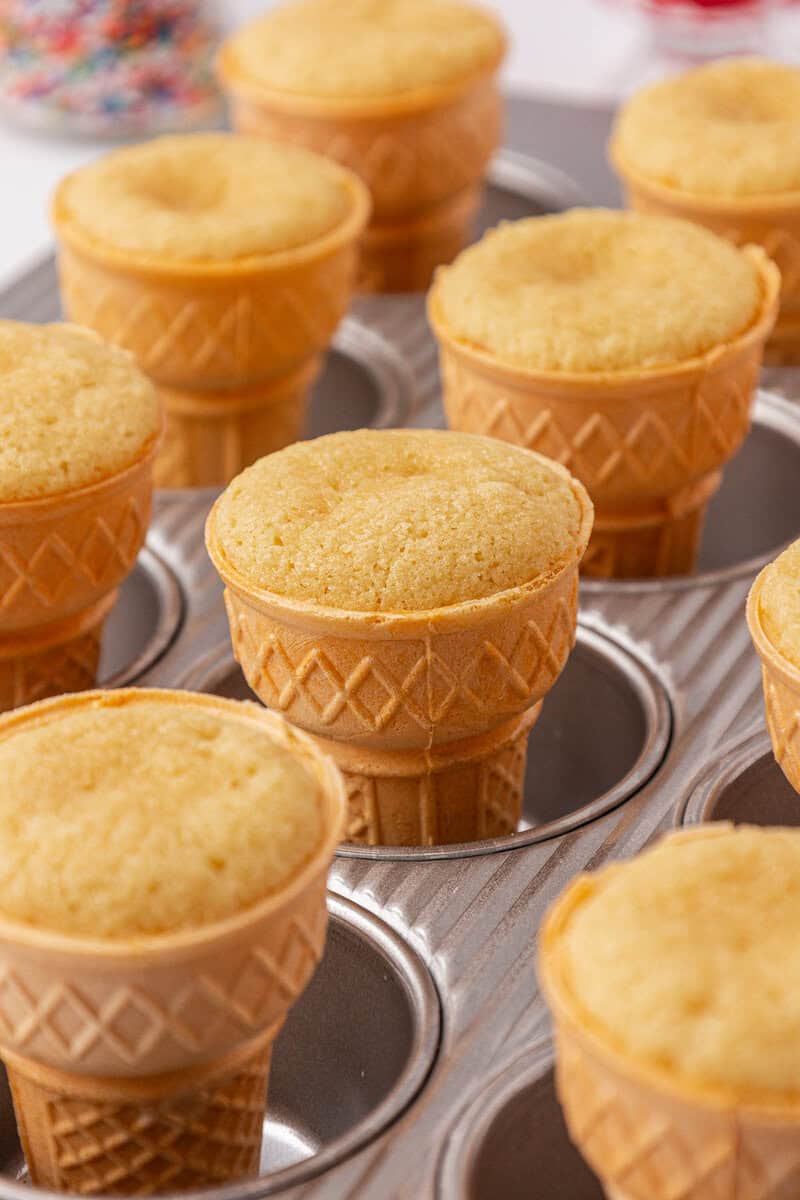 Ice cream cones filled with yellow cake, baked and arranged upright in a muffin tin. The cones have a waffle pattern and the cake tops are golden and slightly domed. Sprinkles are blurred in the background.
