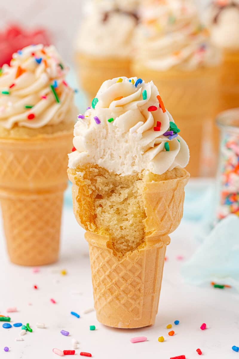 A cupcake baked in an ice cream cone, topped with swirled frosting and colorful sprinkles, has a bite taken out of it. Other frosted cupcake cones and scattered sprinkles are visible in the background.