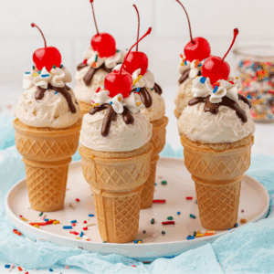 Five ice cream cones topped with vanilla ice cream, whipped cream, chocolate drizzle, rainbow sprinkles, and a cherry on top are arranged on a plate with scattered sprinkles around them.