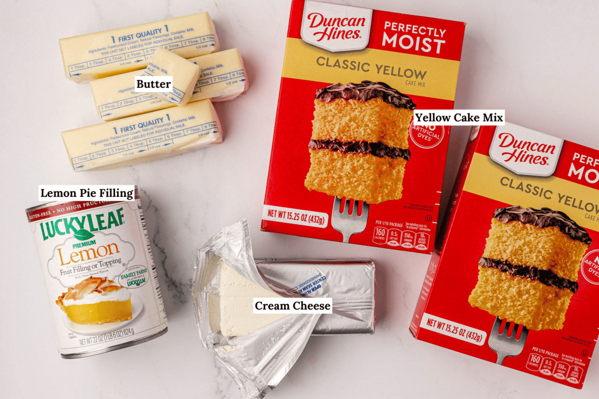 Image showing labeled ingredients for a dessert: three sticks of butter, two boxes of yellow cake mix, a can of lemon pie filling, and a package of cream cheese on a white background.
