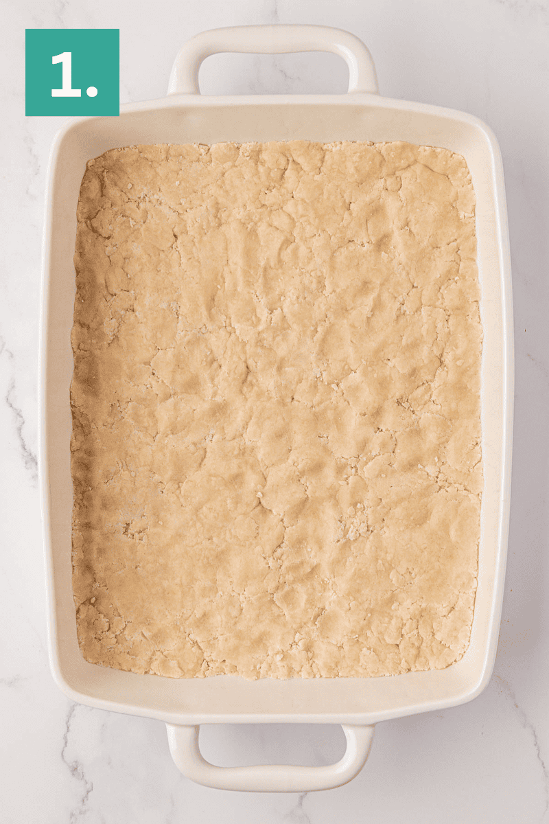 A rectangular white baking dish filled with an even layer of unbaked dough sits on a marble surface. A green square in the top left corner displays the number “1.”.