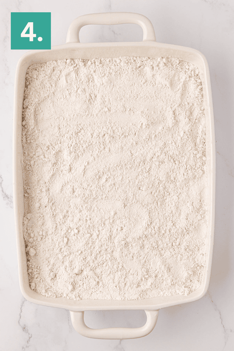 A rectangular white baking dish filled with a layer of flour mixture sits on a white marble surface. A green box in the top left corner displays the number 4.