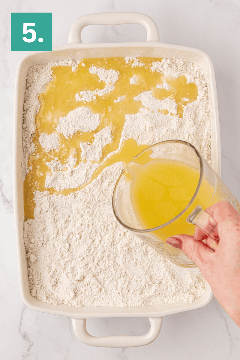 A hand pours melted butter from a measuring cup onto a layer of flour in a white rectangular baking dish. A green box with the number 5 is in the upper left corner.