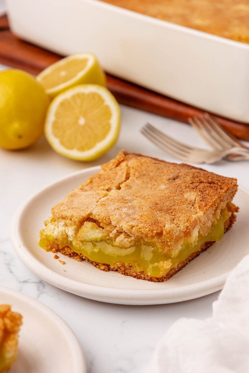 A slice of lemon dessert bar with a golden, crumbly top sits on a plate. In the background are a baking dish, two cut lemons, and two forks on a white marble surface.