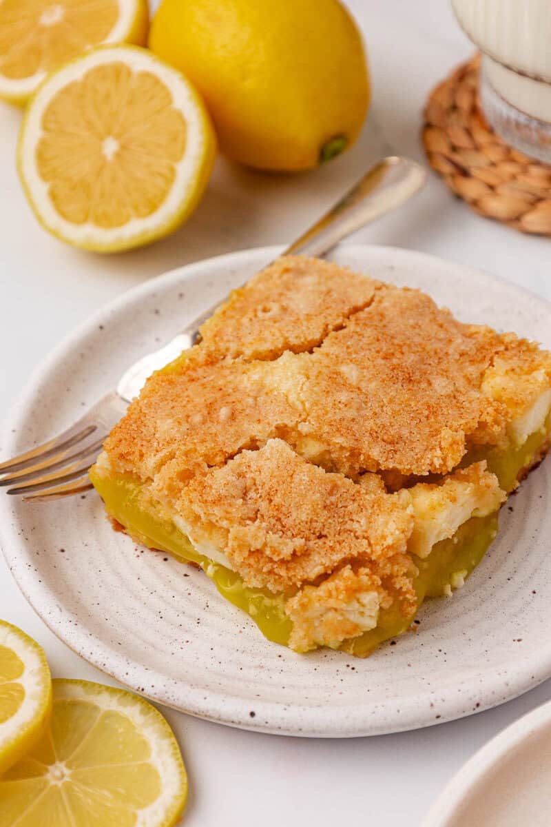 A square slice of lemon bar with a crumbly golden crust sits on a white plate with a fork, surrounded by fresh lemon halves and whole lemons in the background.