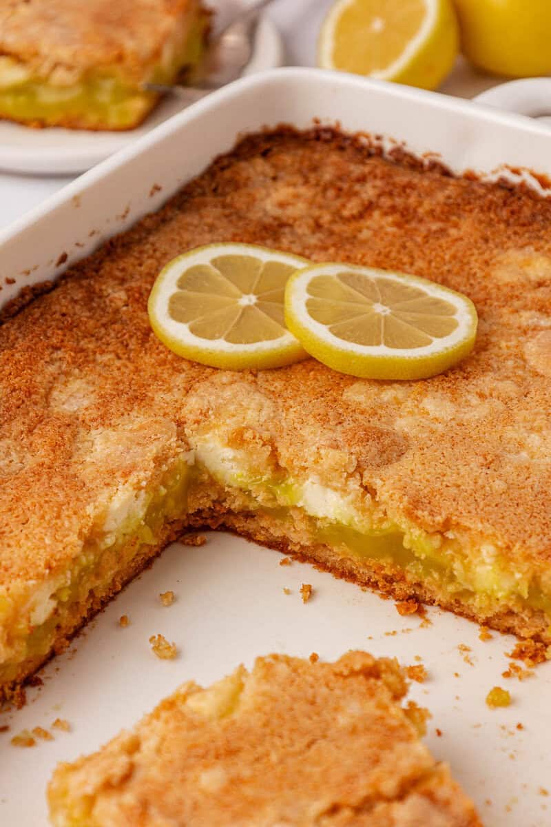 A baked lemon dessert with a golden crust is shown in a white baking dish. Two lemon slices rest on top, and a square piece has been cut out, revealing a gooey lemon filling inside.