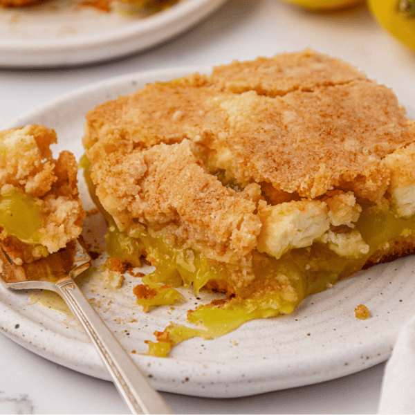 A close-up of a slice of lemon pie with a crumbly golden crust and gooey lemon filling on a white plate; a fork rests beside the pie holding a small bite.