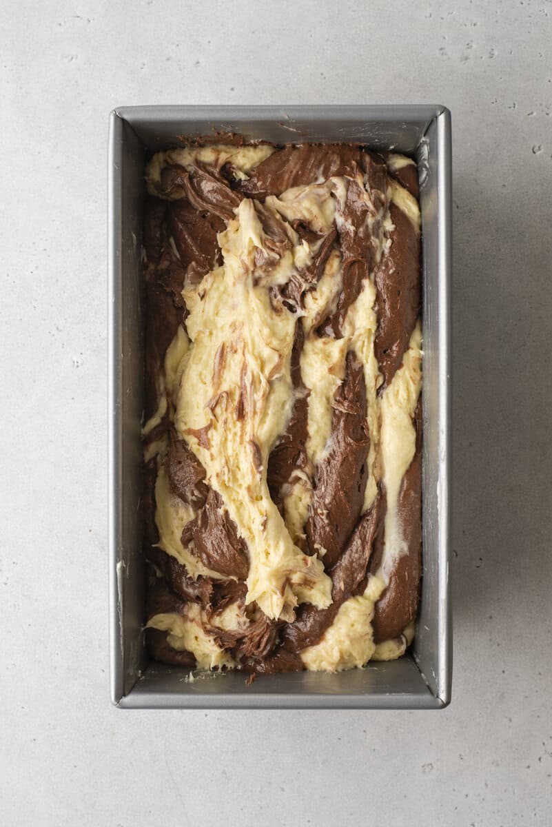 A loaf pan filled with unbaked chocolate and vanilla swirled cake batter sits on a light gray countertop. The batters are marbled together, creating a textured pattern.