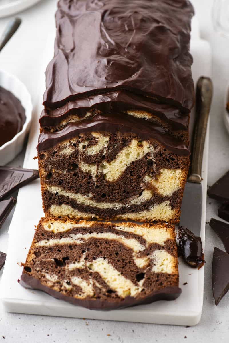 A sliced marble loaf cake with chocolate and vanilla swirls, topped with glossy chocolate icing, displayed on a white cutting board with a knife and pieces of chocolate nearby.