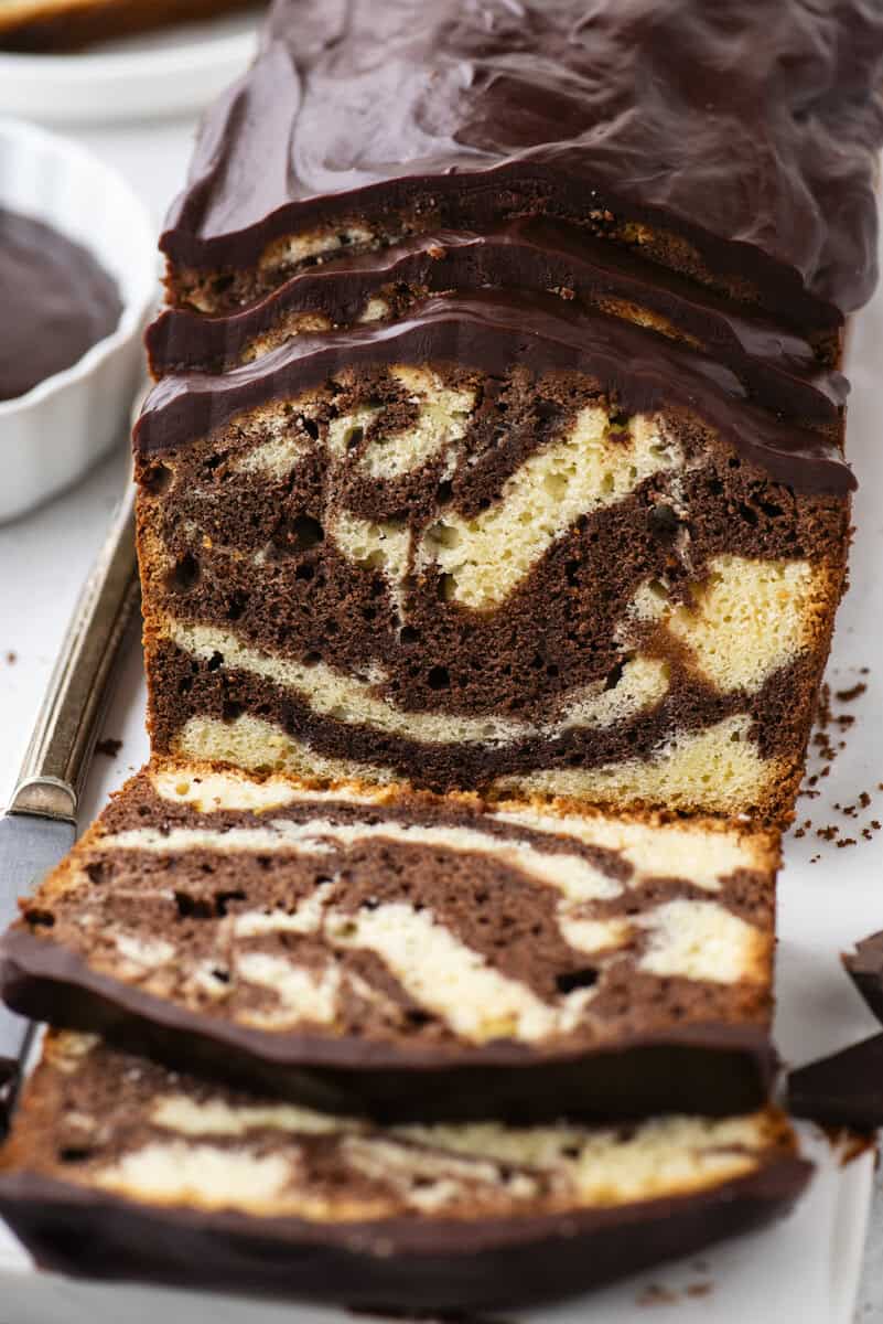 A sliced marble loaf cake with swirls of vanilla and chocolate, topped with a glossy layer of chocolate glaze, is displayed on a white surface with a knife nearby.