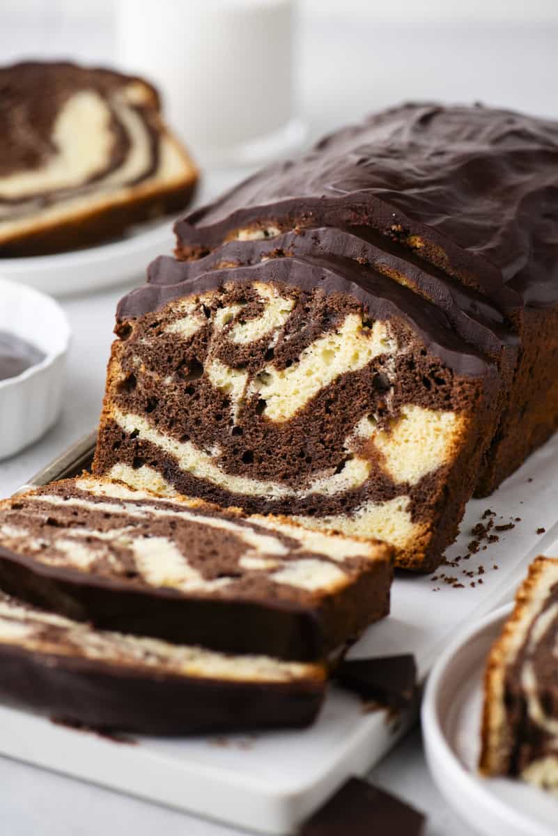 A sliced marble loaf cake with swirls of chocolate and vanilla, topped with a layer of chocolate glaze, sits on a white cutting board next to a glass of milk.