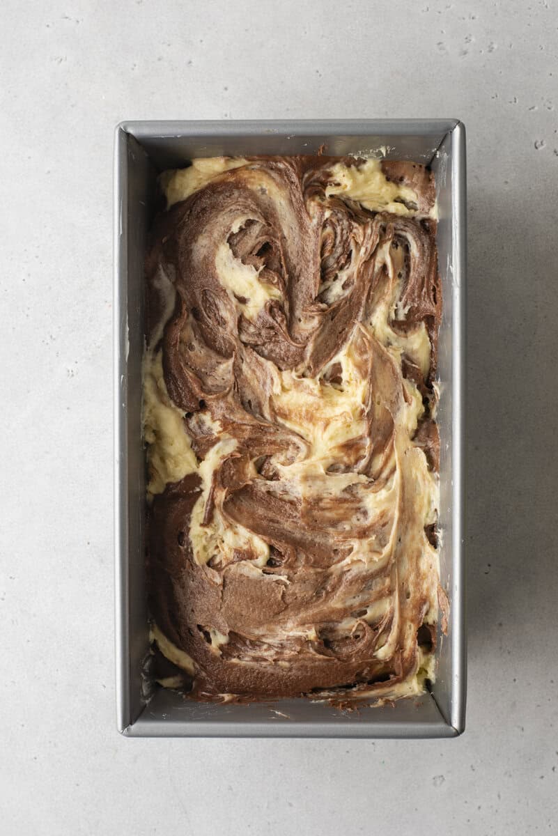 A rectangular metal baking pan filled with unbaked chocolate and vanilla marbled cake batter, creating a swirled pattern, sits on a light gray surface.