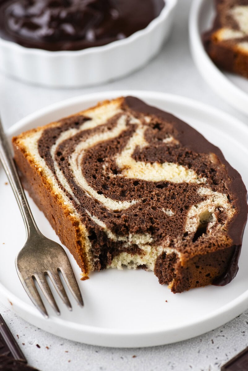 A slice of marble cake with a chocolate glaze is on a white plate next to a fork. The cake has a bite taken out, showing the swirled chocolate and vanilla pattern.
