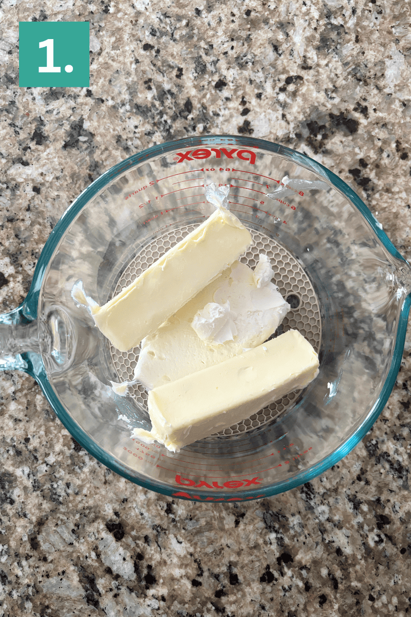 A glass measuring cup with two sticks of butter sits on a granite countertop. A green box with the number 1 appears in the top left corner of the image.