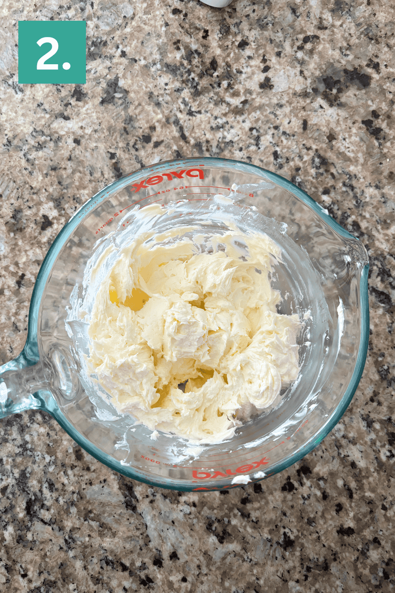 A glass measuring cup with whipped, creamy butter mixture sits on a marbled countertop. A green box with the number 2 is in the top left corner.