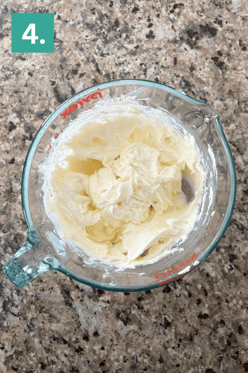 A glass measuring cup filled with whipped cream cheese sits on a speckled granite countertop. A green box in the top left corner displays the number 4.