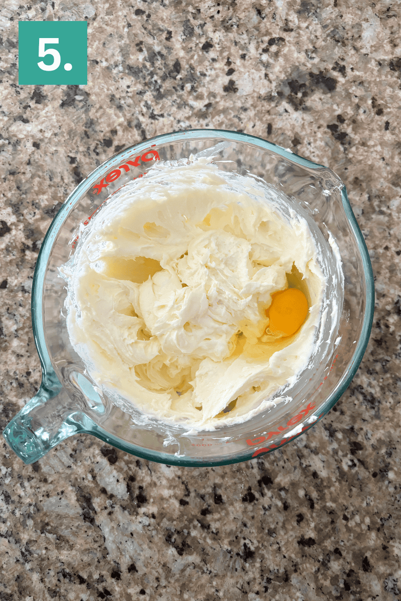 A glass measuring cup on a granite countertop containing creamed butter and sugar with an egg on top. The image is labeled with a green box and the number 5 in the upper left corner.