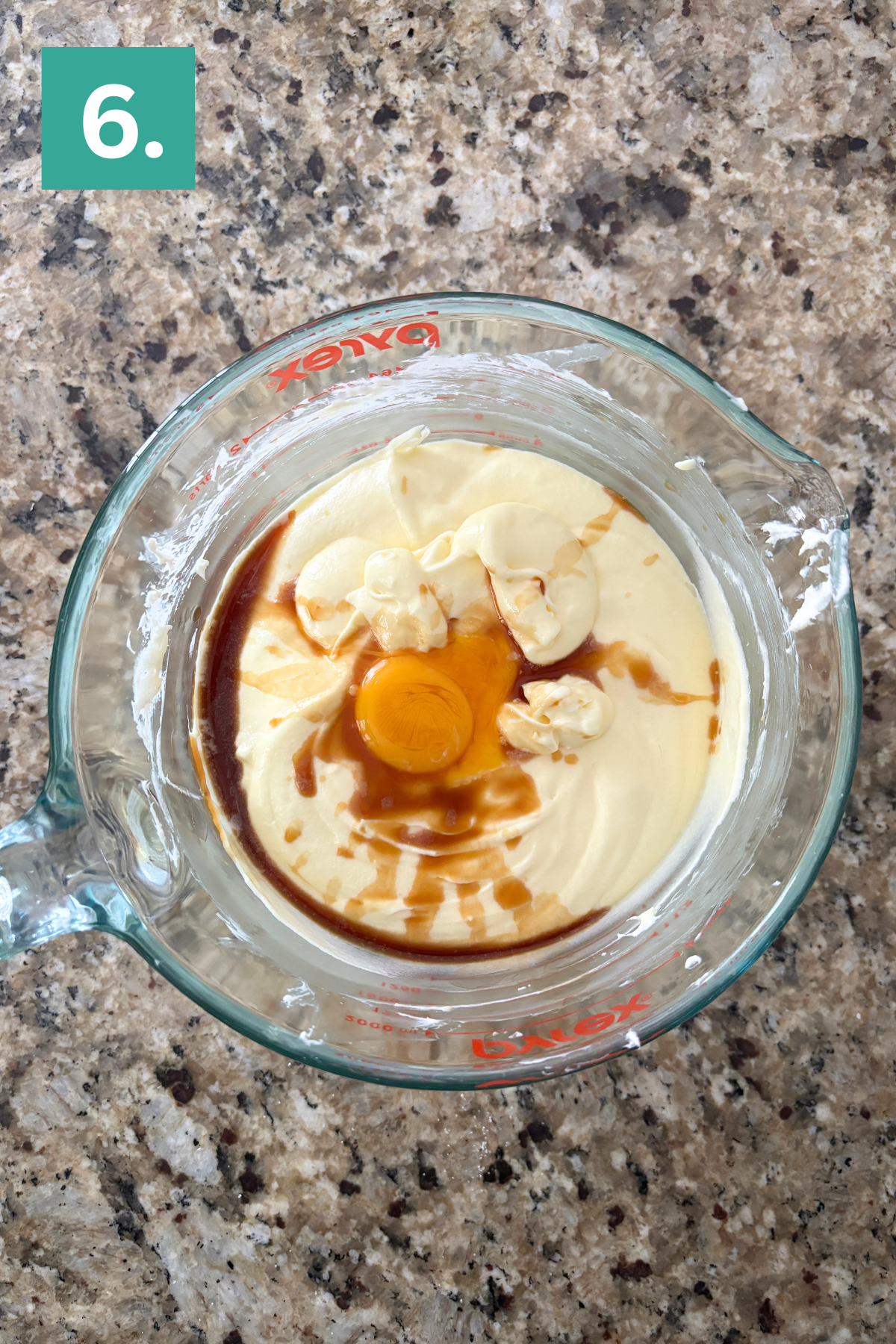 A glass mixing bowl on a granite countertop contains a creamy yellow batter with an egg, vanilla extract, and dollops of butter on top. A green box labeled 6. is in the upper left corner.