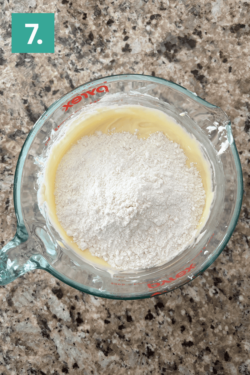 A glass measuring cup with a mixture of flour and wet ingredients sits on a granite countertop. A green box with the number 7 is in the top left corner, indicating a step in a recipe.