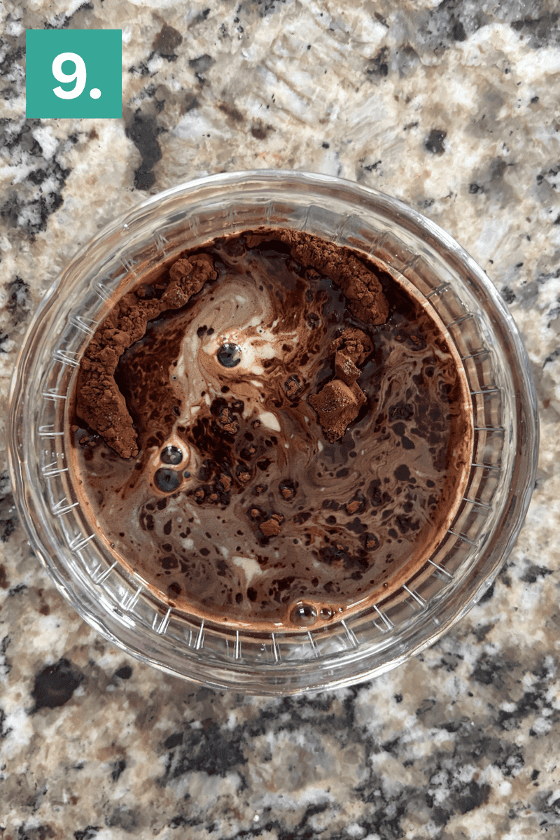 A clear glass bowl with swirled cocoa powder and liquid sits on a speckled granite countertop. A green square with the number 9 is in the top left corner of the image.