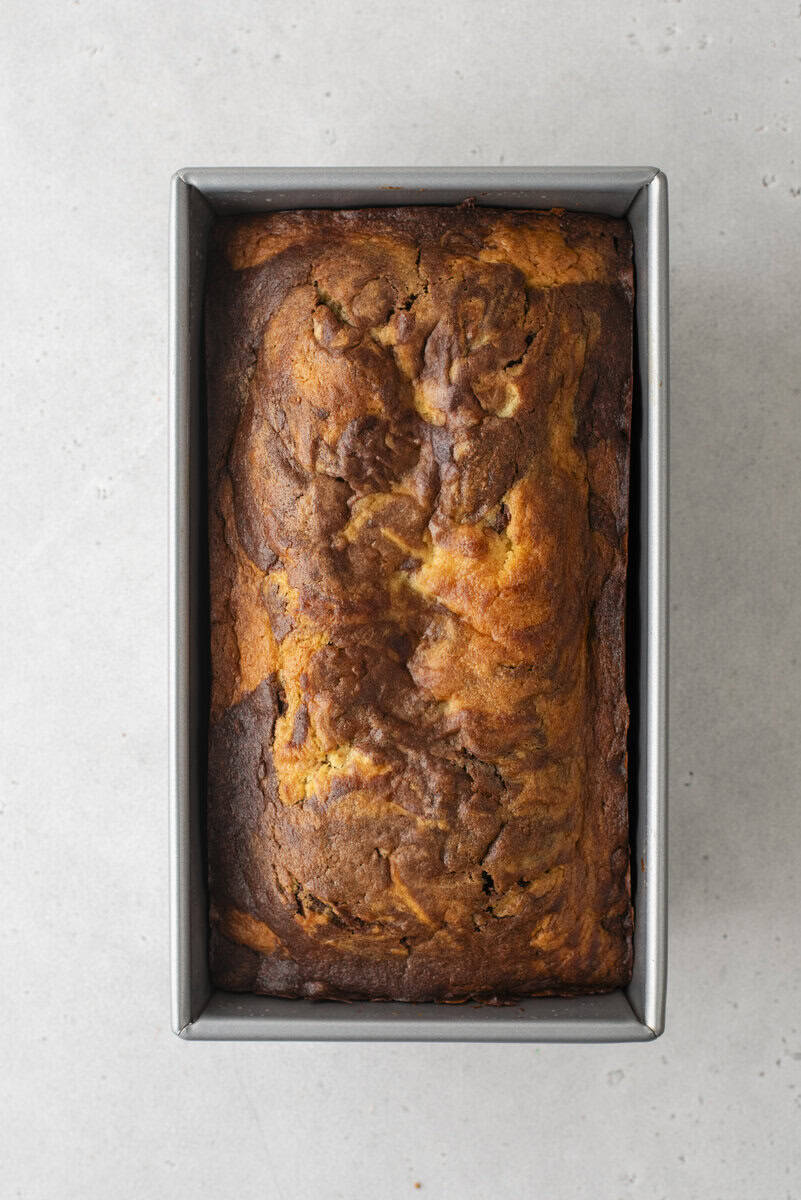 A loaf of banana bread with a golden-brown, slightly cracked top sits in a rectangular metal baking pan on a light gray surface.