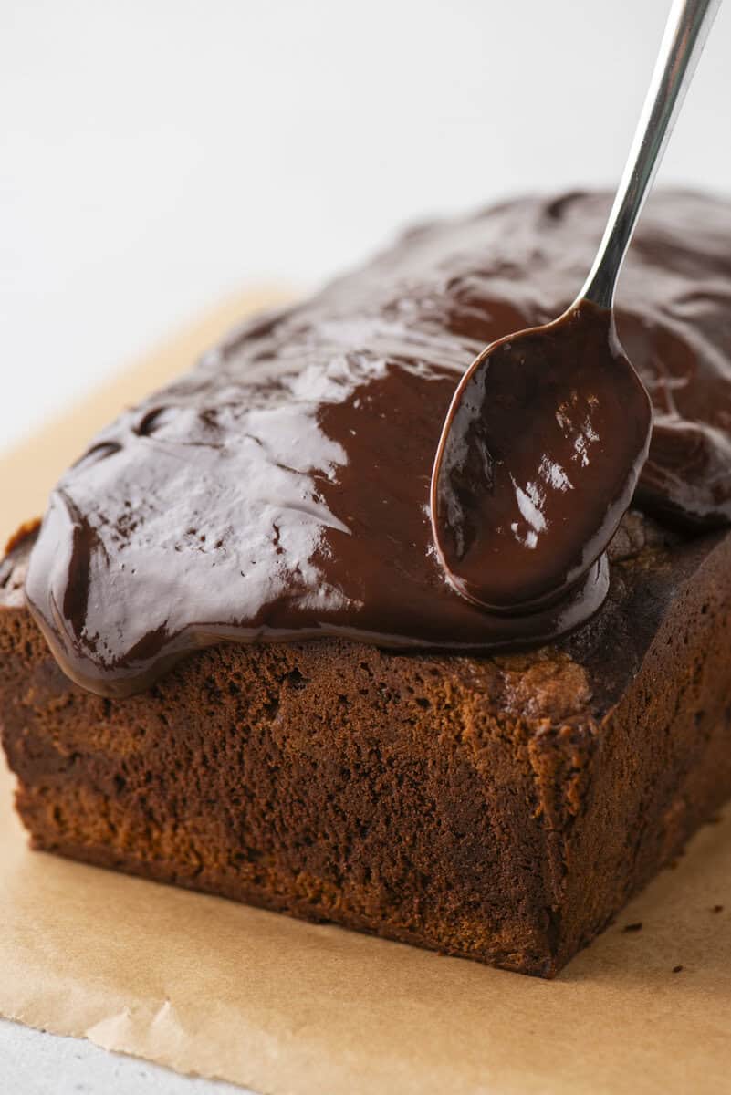 A close-up of a loaf of chocolate cake on parchment paper, with a spoon spreading glossy chocolate icing over the top.