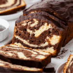 A close-up of a sliced marble loaf cake with swirls of chocolate and vanilla, topped with a glossy layer of chocolate glaze. Slices of the cake are arranged on a white surface.