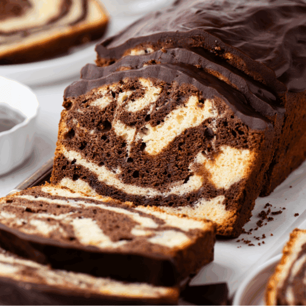 A close-up of a sliced marble loaf cake with swirls of chocolate and vanilla, topped with a glossy layer of chocolate glaze. Slices of the cake are arranged on a white surface.
