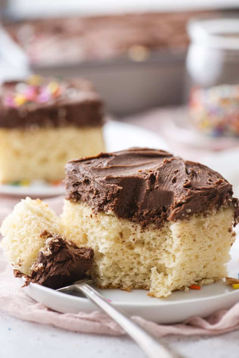 A slice of fluffy yellow cake with thick chocolate frosting sits on a white plate, with a fork holding a bite-sized piece. More cake and a jar of colorful sprinkles are blurred in the background.