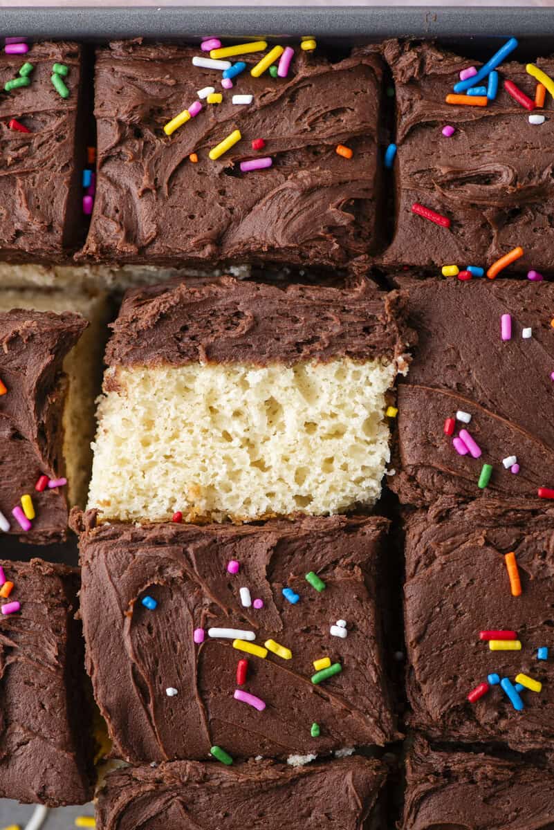 A close-up of a sliced sheet cake with yellow sponge and chocolate frosting, topped with colorful rainbow sprinkles. One slice is partially pulled out from the tray.