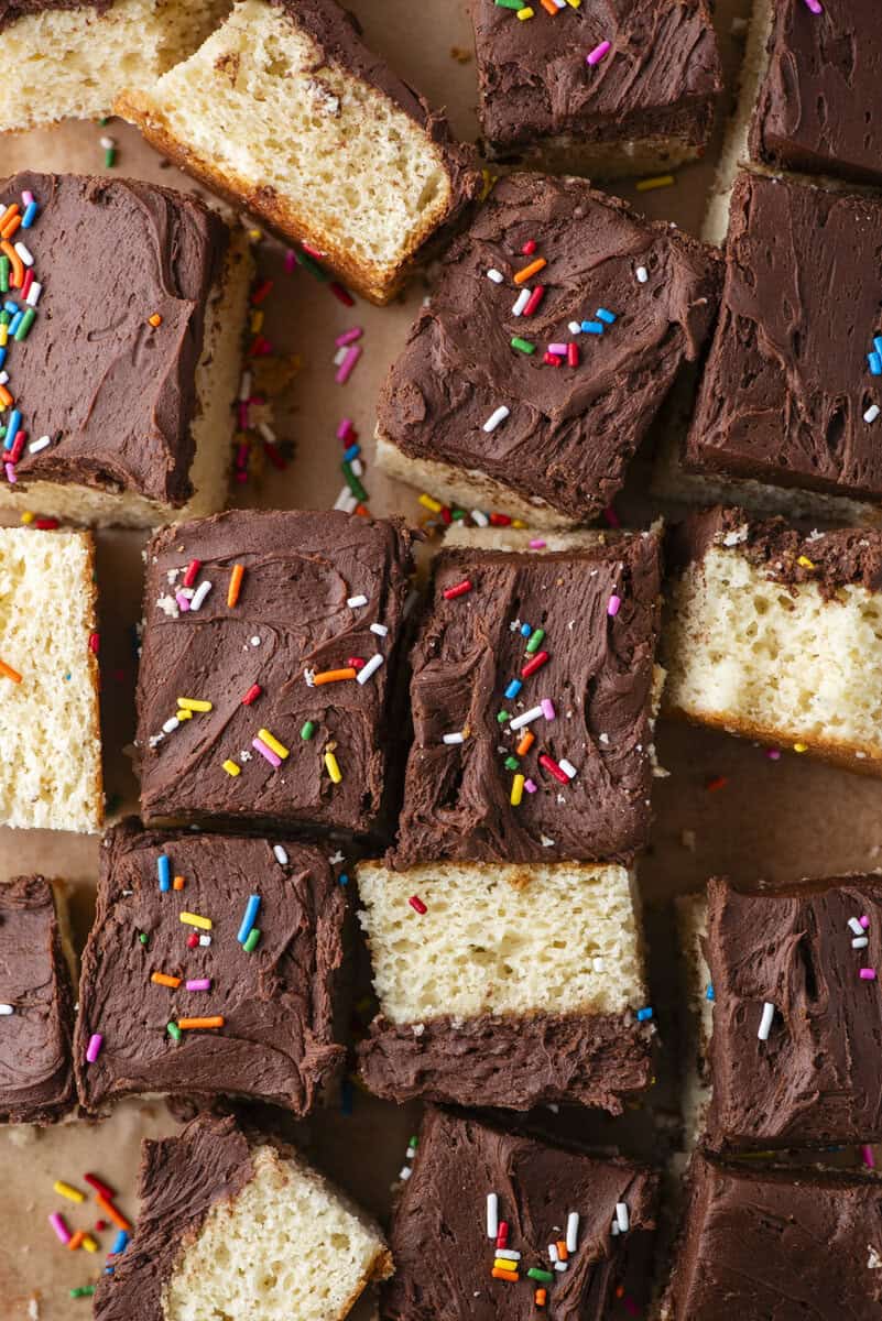 Squares of yellow cake topped with chocolate frosting and colorful rainbow sprinkles are arranged on a brown surface. Some cake pieces are packed closely together, while others are slightly apart.