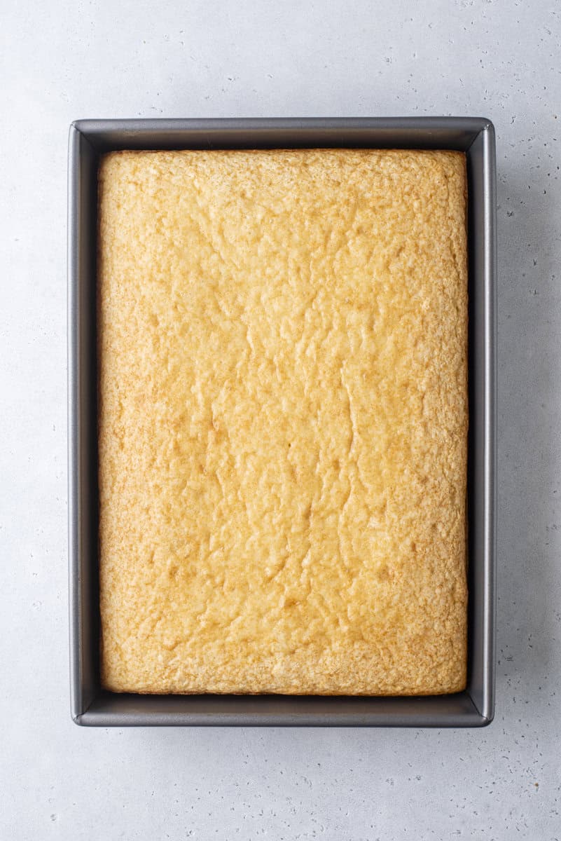 A rectangular, golden-brown sheet cake sits in a dark metal baking pan on a light gray surface. The cake appears plain, without any frosting or decorations.