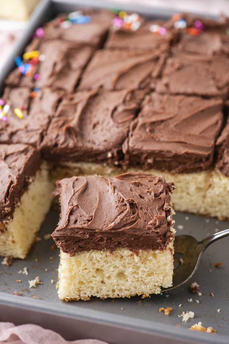 A metal baking pan filled with square pieces of yellow cake topped with chocolate frosting. One frosted square is being lifted out on a spatula, and there are colorful sprinkles on some pieces in the background.