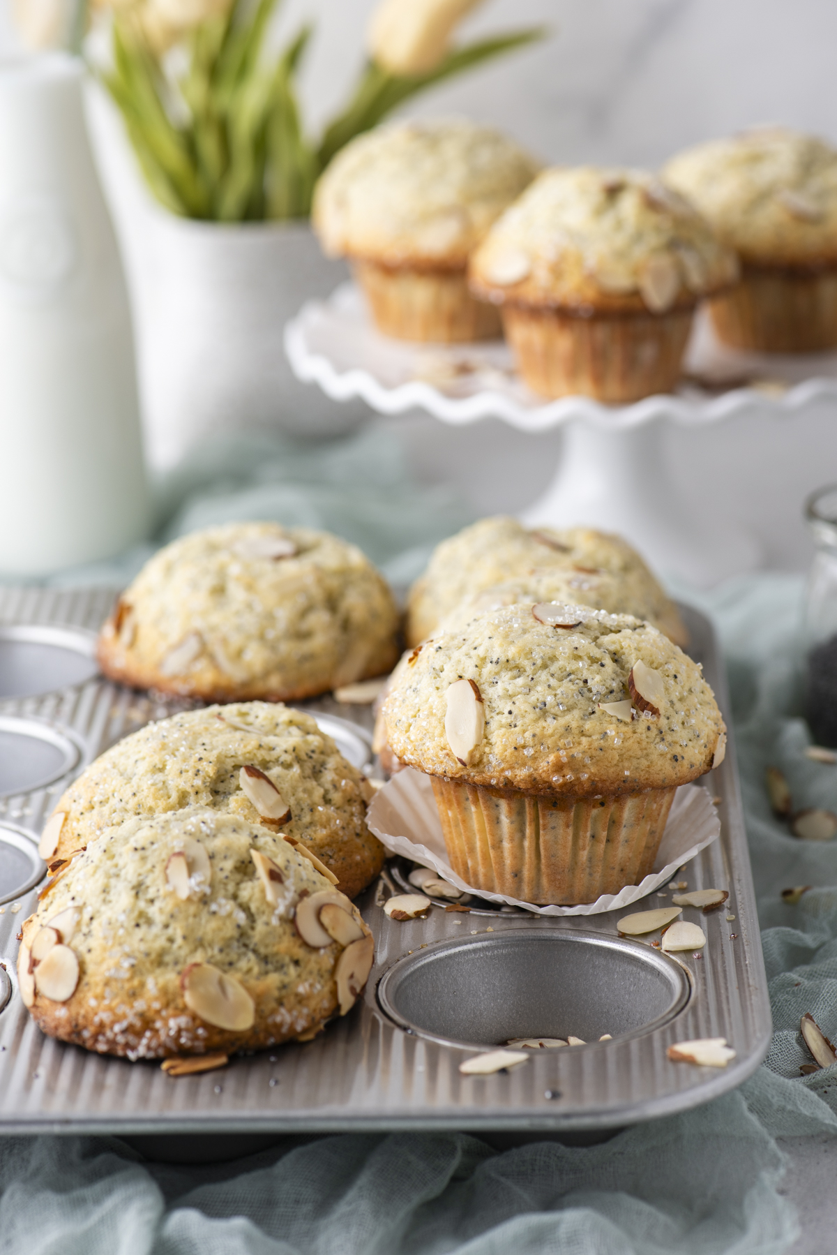 A close-up of freshly baked almond muffins in a muffin tin, with sliced almonds on top. A few muffins are on a stand in the background, and a vase with flowers and a bottle of milk are also visible.