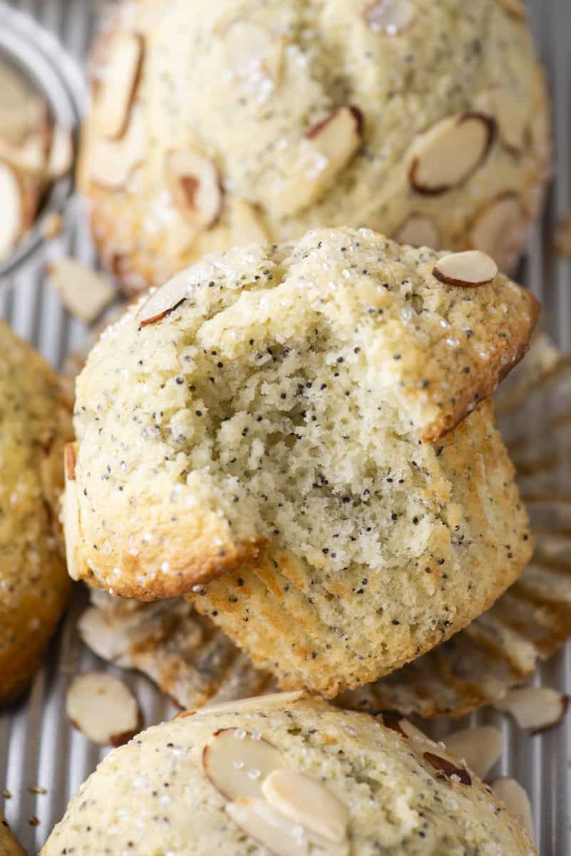 A close-up of poppy seed muffins topped with sliced almonds. One muffin is broken in half, showing its soft, moist texture. The muffins rest on a metal tray with scattered almond slices.