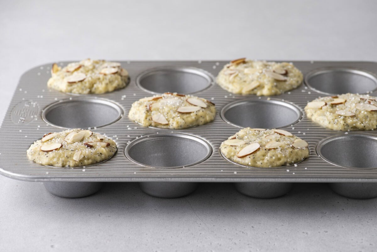 A metal muffin tin holds five unbaked muffins topped with sliced almonds and coarse sugar, while four cups remain empty; the tin sits on a light gray surface.