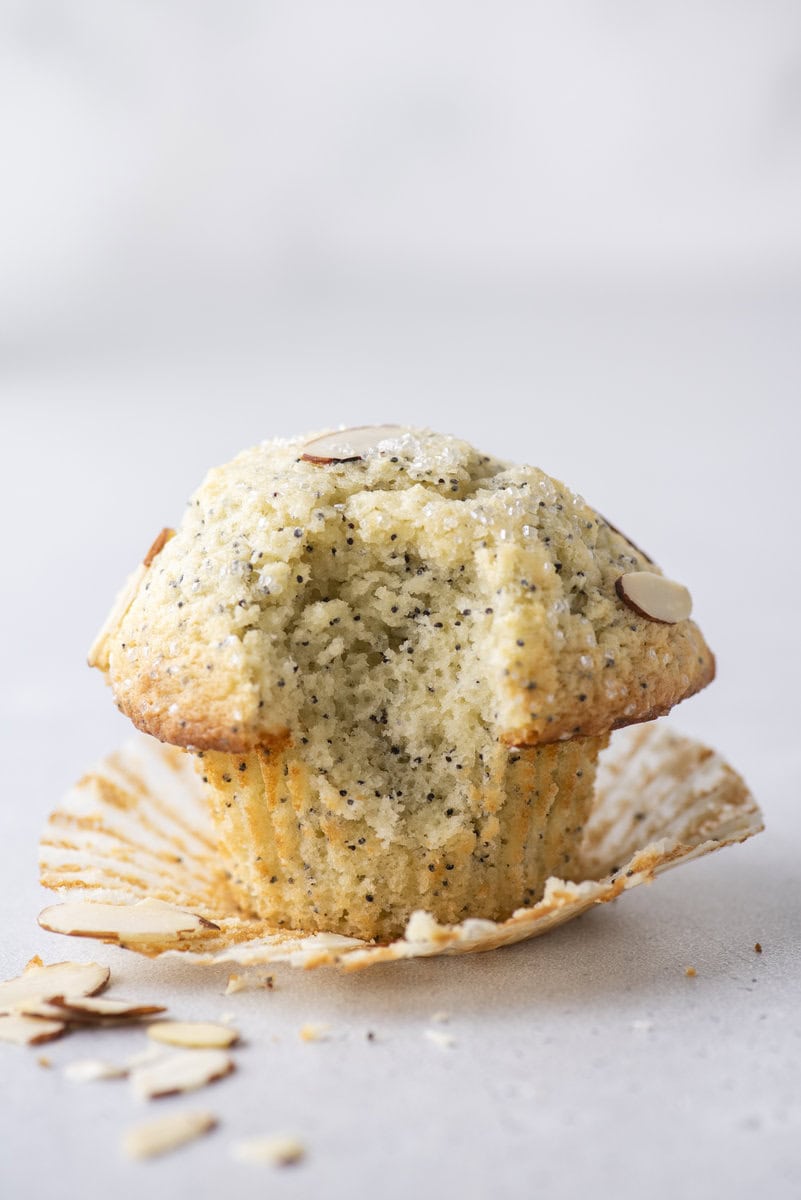 A lemon poppy seed muffin with a bite taken out, sitting on an open muffin wrapper. Sliced almonds and crumbs are scattered nearby on a light surface.